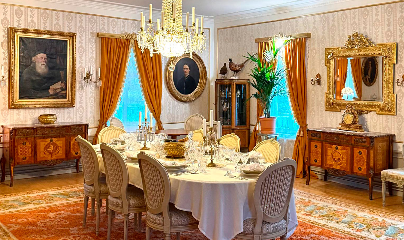 Kuva kartanon ruokapöydästä, joka on katettu. Seinillä on tauluja ja lattialla pöydän alla suuri matto.