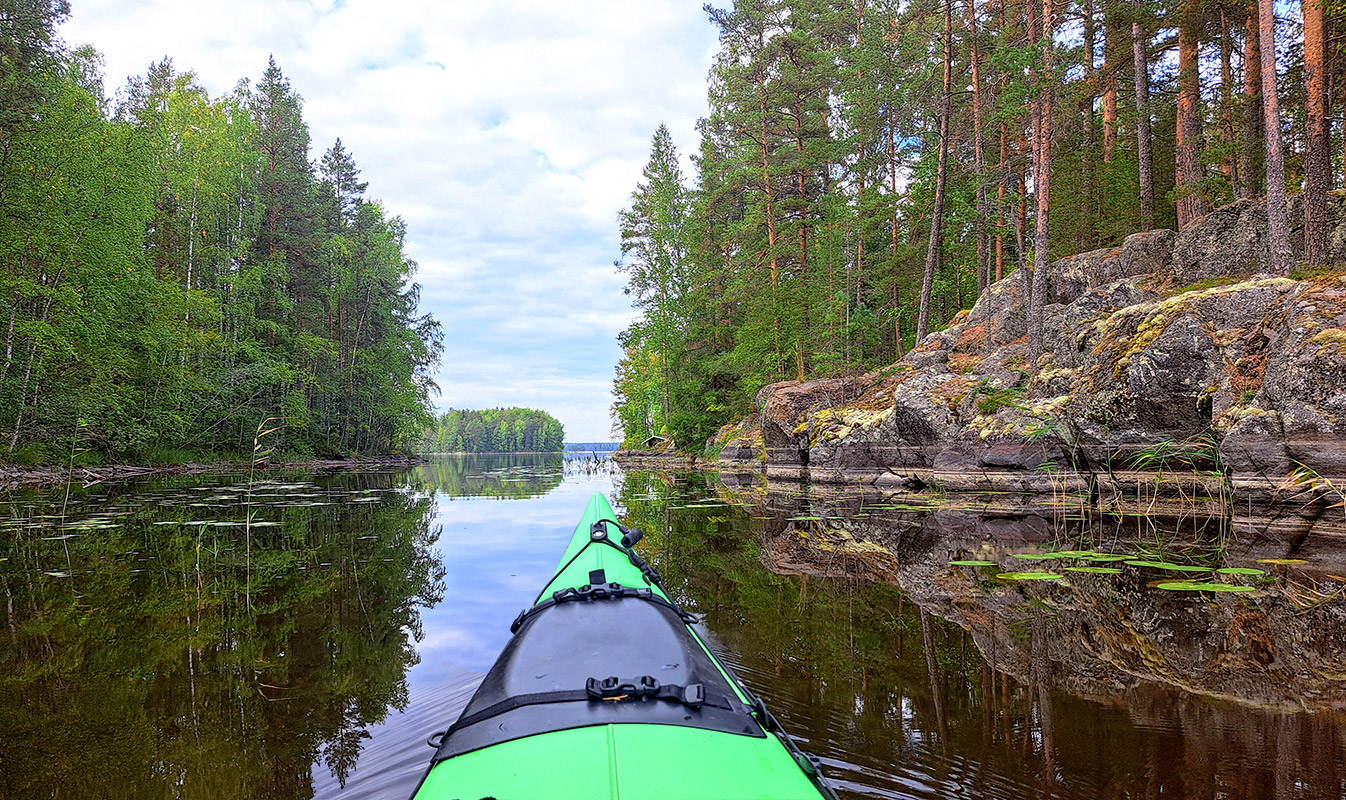 Aittosalmen kalliot ja peilityyni järvi kallioisen rannan vieressä, kajakki lipuu veden pinnalla.