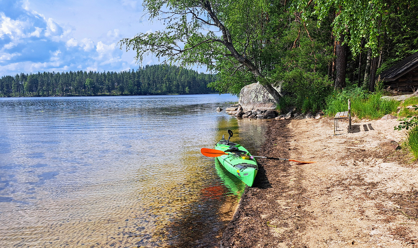 Kajakki hiekkarannalla, tyyni järvi ja metsäinen horisontti.