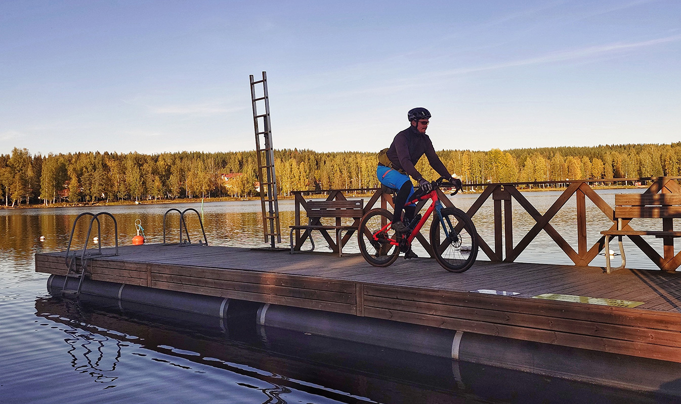 Pyöräilijä laiturilla järven äärellä, metsä värjäytynyt oranssiksi auringonvalossa.