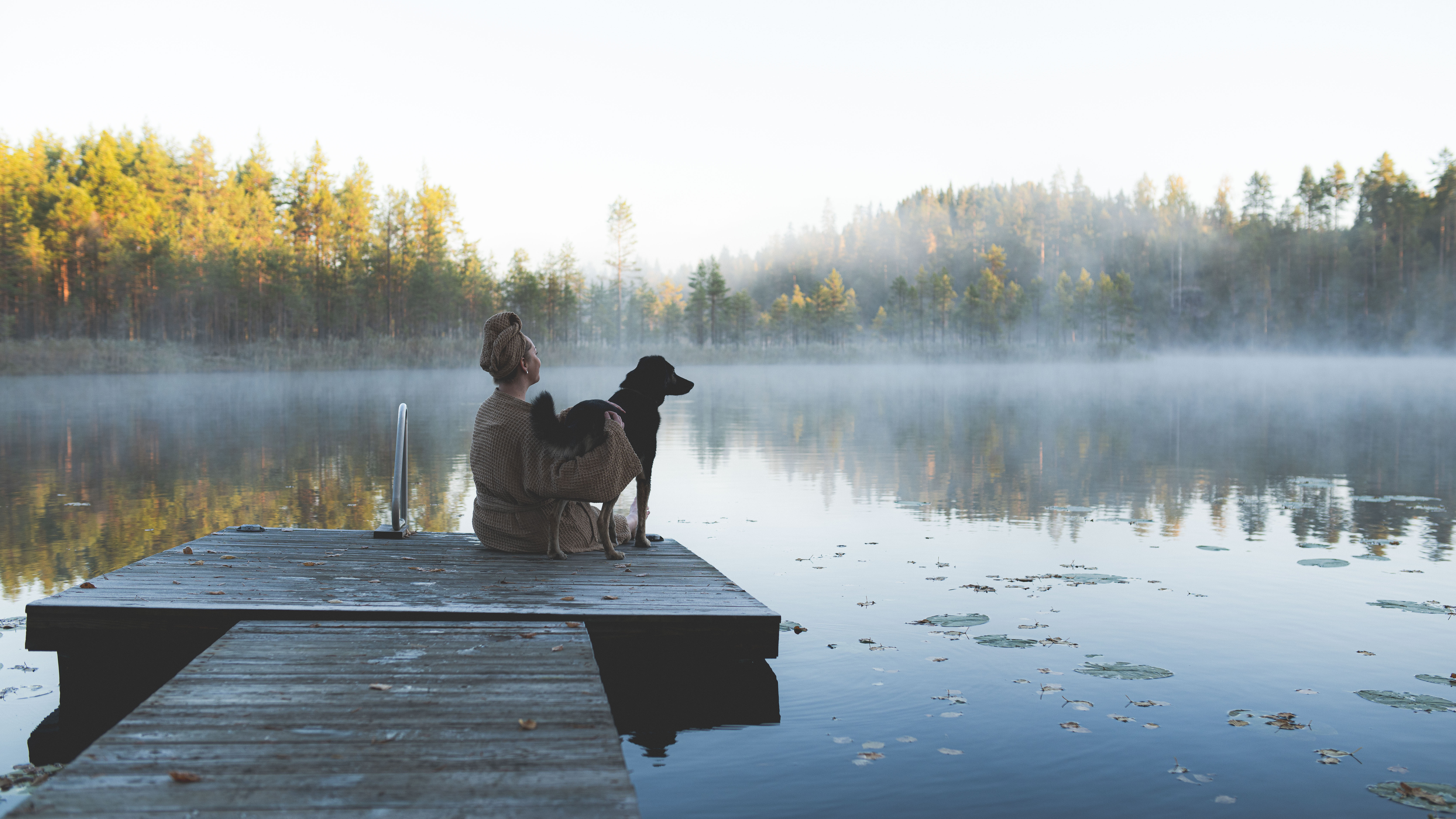 Henkilö istuu laiturilla koiran kanssa, taustalla usvainen järvimaisema.