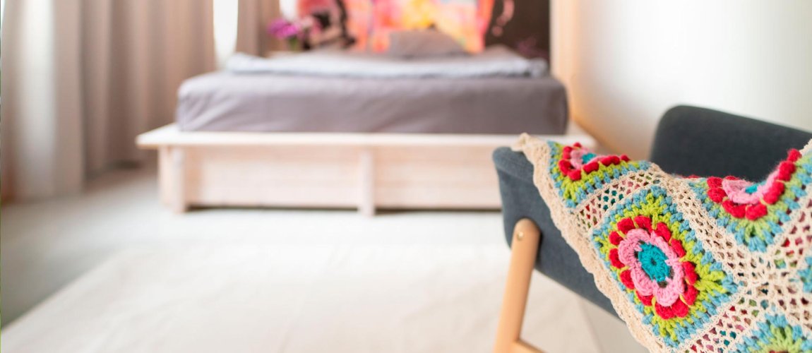 Nojatuolilla viltti, taustalla sänky ja värikäs tapetti pehmeästi sumentuneena.