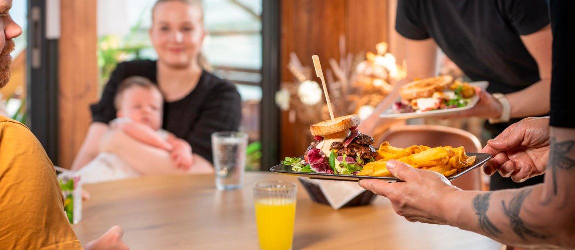 Tarjoilija ojentaa täytetyn toastin, taustalla aikuinen, jolla vauva sylissä.