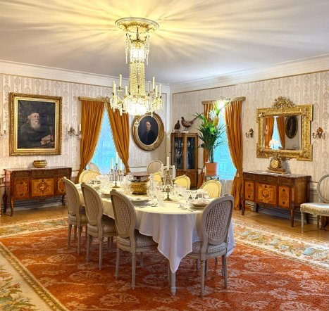 Kuva kartanon ruokapöydästä, joka on katettu. Seinillä on tauluja ja lattialla pöydän alla suuri matto.