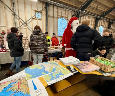 Kuva junatallista, jossa on myyntipöytiä ja ihmisiä. Etualalla hankkeen piste, jossa karttoja ja infoa Äänekoskesta.